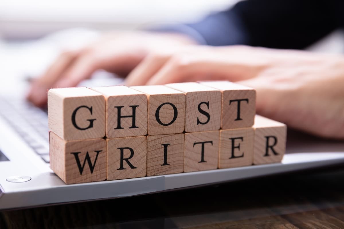 Blocos de madeira com letras compondo a palavra ghost-writer em cima do teclado de um notebook em que um homem digita.